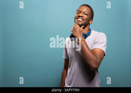 Confident young man wearing a wireless headset around his neck and holding his chin in front of an isolated backdrop. African american guy with headphones, touching his jaw and smiling at camera. Stock Photo