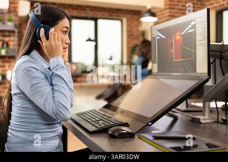 Female designer puts on her headphones and works on digital monitors, improving visual designs in a modern studio. Asian woman touching her earphones, and looking at her designs on computer screen. Stock Photo