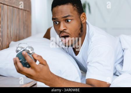 Shocked Black Guy Looking At Alarm-Clock Waking Up Oversleeping Indoors Stock Photo
