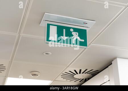 Ceiling mounted LED emergency fire exit sign with arrow pointing towards the fire door in a UK office, with a smoke alarm behind. Concept: fire safety Stock Photo