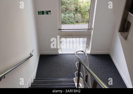 An empty carpeted staircase in a UK office building with a fire exit sign. Concept: fire safety, empty office, office rent, office premises Stock Photo
