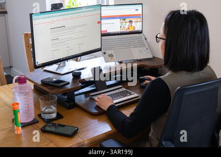 Woman in her thirties wearing work clothes & sitting at a dining room table at home and working using dual computer screens, UK. Concept: home office Stock Photo
