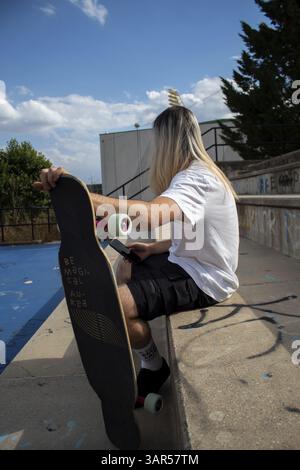 Unrecognizable hipster with long hair with wooden longboard sitting on the steps and chatting in the mobile phone. Concept of leisure activity. Outdoo Stock Photo