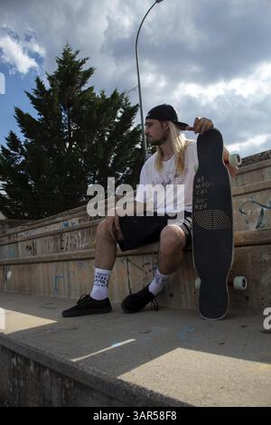 Vertical portrait of a young man with long hair and a cap sitting in the skatepark holding his skateboard and looking at the horizon Stock Photo