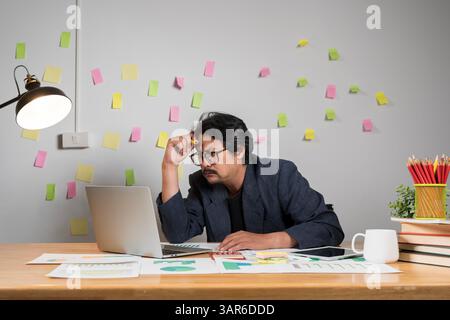 A man with messy hair sits in a chair, intently working on his laptop. A professional man wearing glasses is intently working on a laptop in a cozy, s Stock Photo