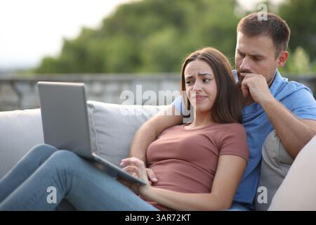Disappointed couple checking laptop content sitting in a couch in a terrace Stock Photo
