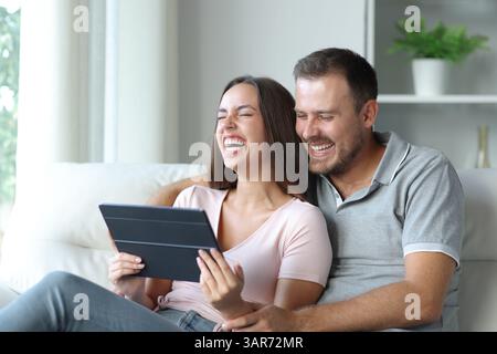 Funny couple watching media content on tablet and laughing loud sitting on a couch at home Stock Photo