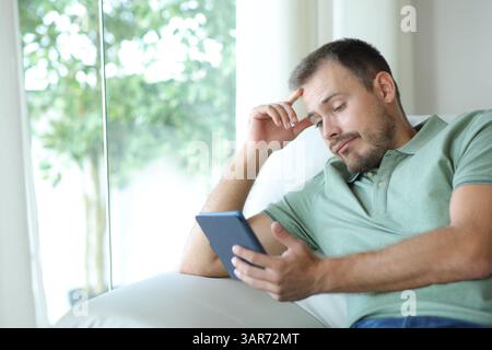 Disappointed man reading a nobel in an ebook sitting on a couch at home Stock Photo