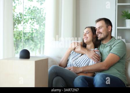Happy couple enjoying looking through a window at home beside a smart speaker Stock Photo