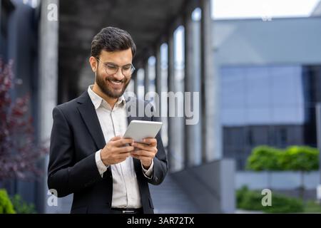 A smiling businessman in a suit works on his tablet while standing outside a modern office building on a sunny day. Stock Photo