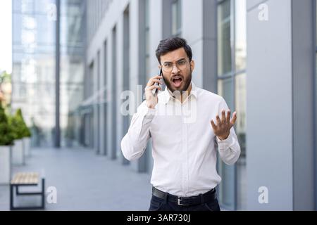 Anxious man is on the phone, expressing frustration with an open mouth and raised hand, outdoors. Stock Photo