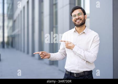 A smiling man with glasses stands outside, pointing towards something. He is wearing a white shirt and looking directly at the viewer. The background has a modern building. Stock Photo