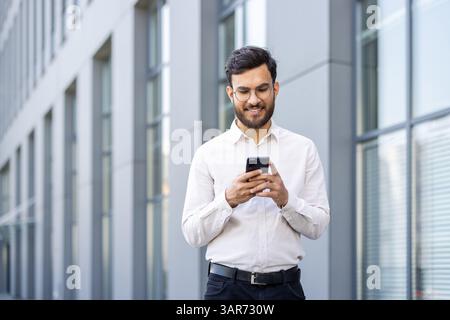A smiling man in glasses checks his smartphone outside a modern building, looking pleased while texting. The sunny day adds to the pleasant scene. Stock Photo