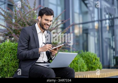 A young businessman in a suit works outdoors, utilizing a laptop, notepad, and earphones, in a modern business district. Stock Photo
