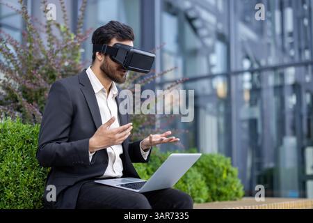 A businessman wearing vr glasses with a laptop is gesturing while sitting outside, modern technology concept. Stock Photo
