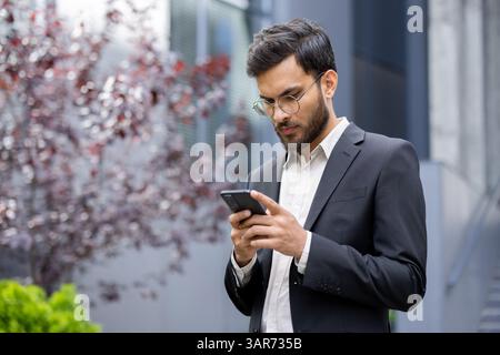 A stylish businessman in a suit uses his smartphone outdoors, surrounded by modern architecture and a blurred background. Stock Photo