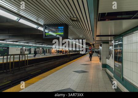 Wide view of Bloor subway station in Toronto showing a train arriving, passengers on platform, and signage under fluorescent lighting. Stock Photo