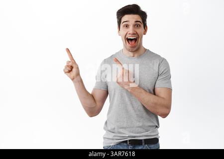 Excited happy guy points aside and laughs. Joyful man shows advertisement on left side, stands over white background. Stock Photo