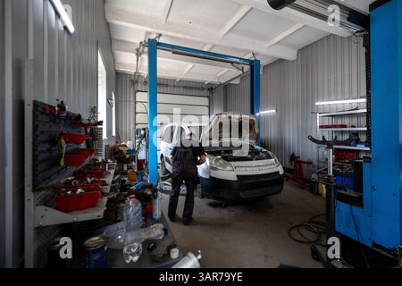 A mechanic works on a white commercial van with its hood open inside a busy auto repair shop equipped with a vehicle lift and various tools Stock Photo