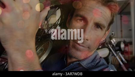 Image of flickering lights over caucasian male mechanic holding motorcycle wheel Stock Photo