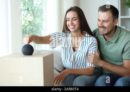 Happy couple starting new smart speaker sitting on a couch at home Stock Photo