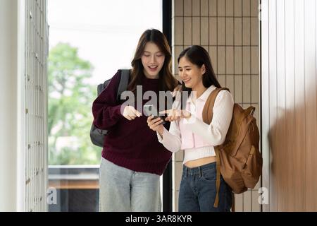 Friendship and College Life. Two students sharing a moment while checking their phones in a school hallway. Stock Photo