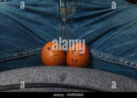 Two brown eggs with drawn faces sit on a person's lap. One egg appears sad while the other looks happy, creating a humorous scene in casual attire. Stock Photo