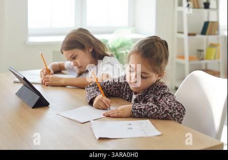 Two children of different ages sitting at table, writing with pencils and using tablet for learning. Stock Photo
