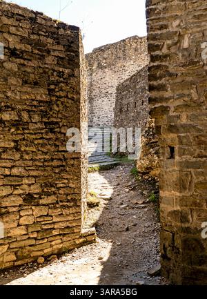 A narrow pathway between ancient stone walls, leading to a staircase in a historical site. The sunlight casts shadows on the rugged surfaces, highligh Stock Photo