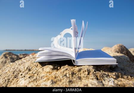 An open book on the beach on a sunny day. A wooden heart as a bookmark. Digital detox, book lovers on vacation Stock Photo