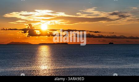 A serene sunset over the ocean, with the sun setting behind clouds and reflecting on the water. Silhouettes of distant islands and a ship are visible< Stock Photo