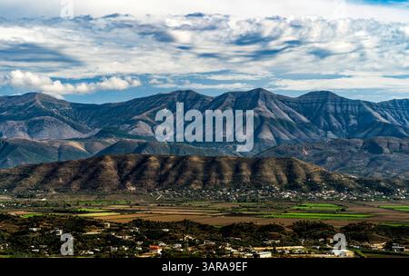 A panoramic view of rugged mountains under a partly cloudy sky, with green fields and small settlements in the foreground. The landscape showcases a m Stock Photo