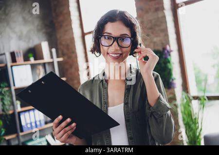Young professional woman with glasses and headset smiling in a casual office, holding a clipboard Stock Photo