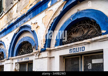 A close-up view of a weathered building facade featuring arched windows with decorative ironwork. The paint is peeling, revealing layers of blue and w Stock Photo