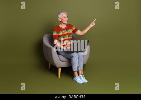Senior Woman Sitting Comfortably With Laptop on Green Background Pointing Upwards Stock Photo