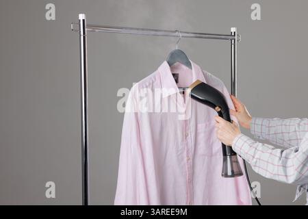Woman steaming pink shirt on hanger using handheld garment steamer to remove wrinkles, home laundry care. Stock Photo
