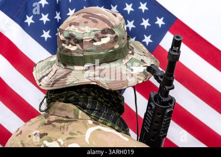 A soldier in camouflage uniform and panama hat with an automatic rifle on his shoulder against the background of the American flag., Concept: military Stock Photo
