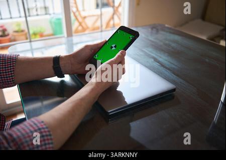 Man interacts with a smartphone displaying a green screen, seated at a table indoors. Nearby, a sleek laptop rests on the desk, depicting a workspace Stock Photo