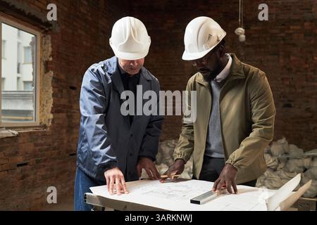 Two biracial builders with helmets on heads having discussion about renovation while concentrated on layout Stock Photo