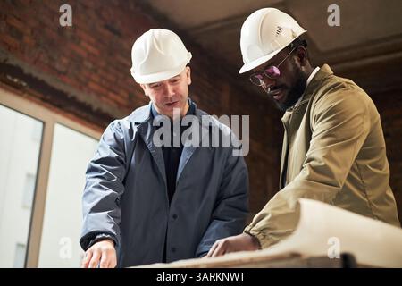 Low angle shot of biracial foremen wearing white helmet having discussion while looking at blueprint on paper Stock Photo