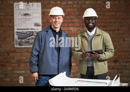 Portrait of African American designer of interior with tablet in hand and Caucasian foreman smiling at camera while posing for photo at contruction site Stock Photo