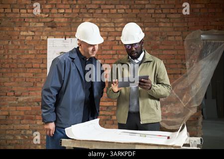 African American designer of interior showing layout plan of IT office on tablet to Caucasian foreman dressed in uniform Stock Photo