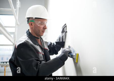 Medium shot of serious Caucasian man wearing hardhat and protective glasses using smooth paste to putty holes on wall Stock Photo