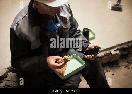 High angle shot of tired laborer sitting on broken floor enjoying sandwich and holding smartphone Stock Photo