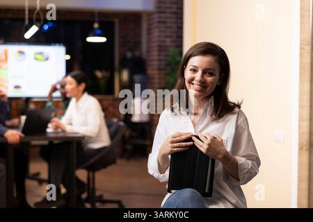 White businesswoman in white shirt smiles confidently while holding a tablet in a modern startup office. The late night workplace, brick walls and multiethnic team highlight productivity. Stock Photo