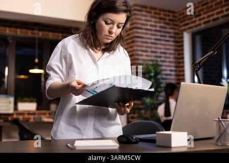 Professional caucasian woman stands at her desk, holding a clipboard and flipping through documents. She carefully reviews company research data in a modern brick walled office late at night. Stock Photo