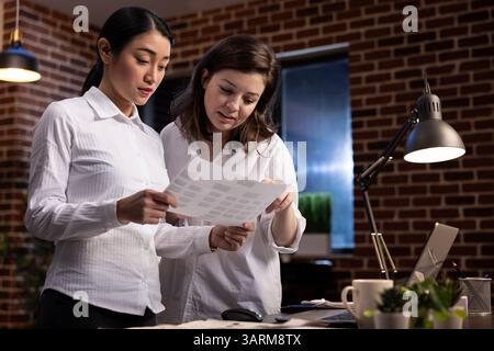 Two female colleagues engage in a late night meeting, discussing documents and sharing ideas. Asian and caucasian women standing and working together to strategize on company branding. Stock Photo