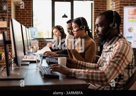 Team of diverse telemarketers sits at their desks, assisting remote clients and managing business inquiries. Headsets, coffee, and computers highlight the dynamic customer support workspace. Stock Photo