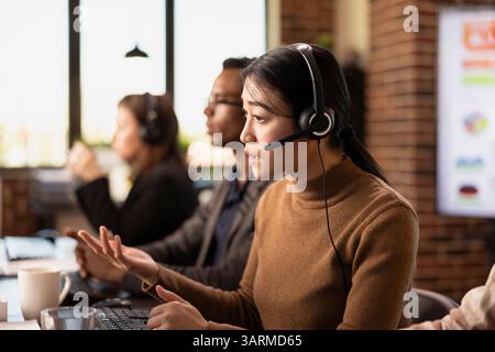 Asian businesswoman wearing wired headphones and providing assistance to remote clients. Serious female call operator with headset sitting at desk and handling customer inquiries. Stock Photo