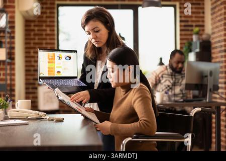 Businesswomen researching marketing trends, checking reports on digital device and clipboard. Caucasian financial advisor with laptop, explains investment opportunities to asian coworker in wheelchair Stock Photo